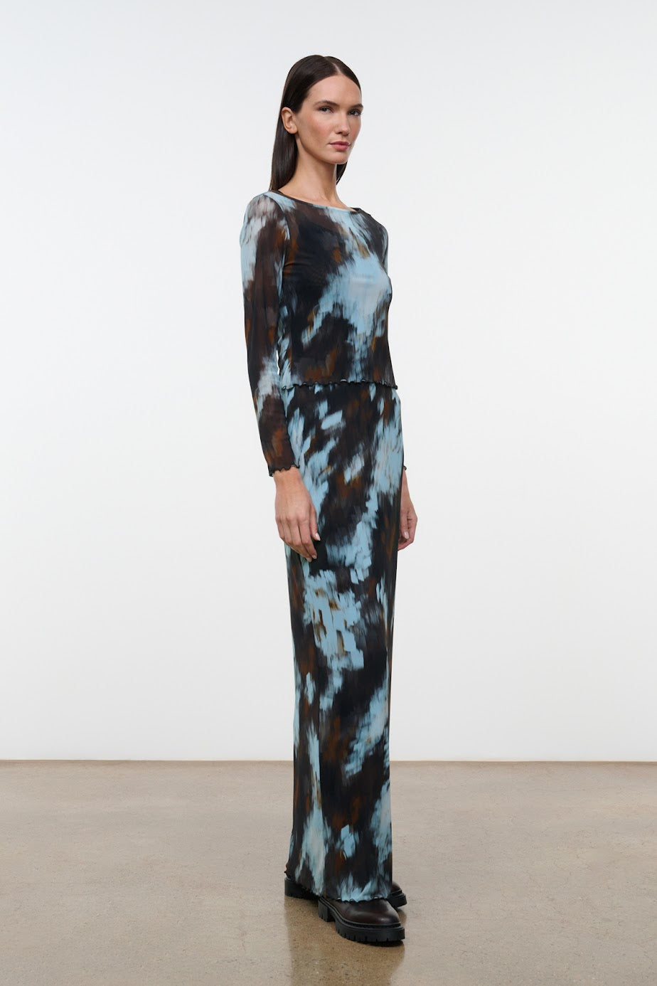 A woman modeling a long-sleeve maxi dress with a blue, brown, and black abstract tie-dye pattern, standing in a minimalist studio with a white wall and light concrete floor.