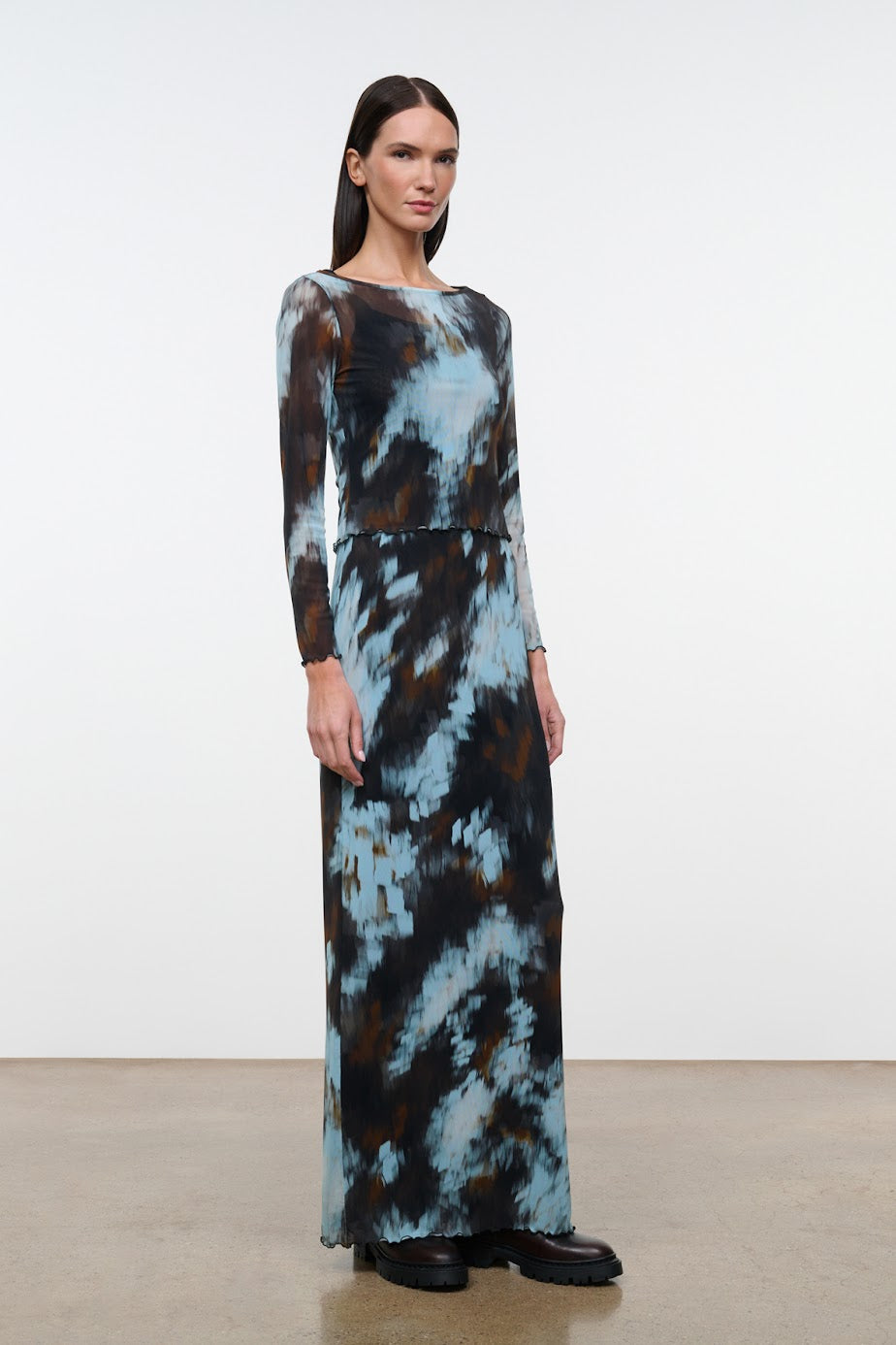 A woman modeling a long-sleeve maxi dress with a blue, brown, and black abstract tie-dye pattern, standing against a plain white studio background.