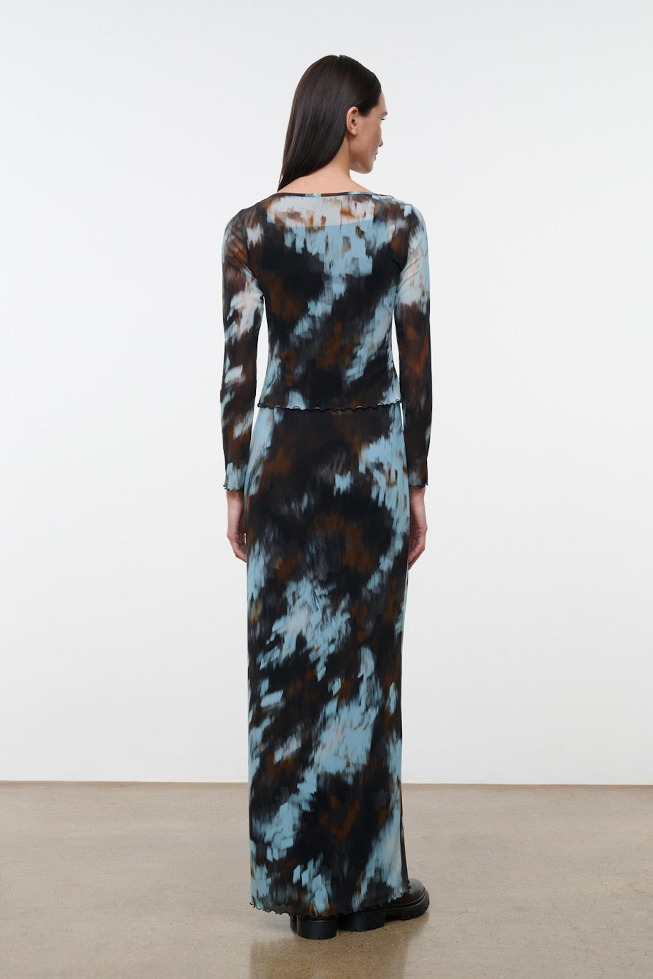 A woman viewed from the back wearing a long-sleeve maxi dress with a blue, brown, and black abstract tie-dye pattern, standing against a plain white studio wall.
