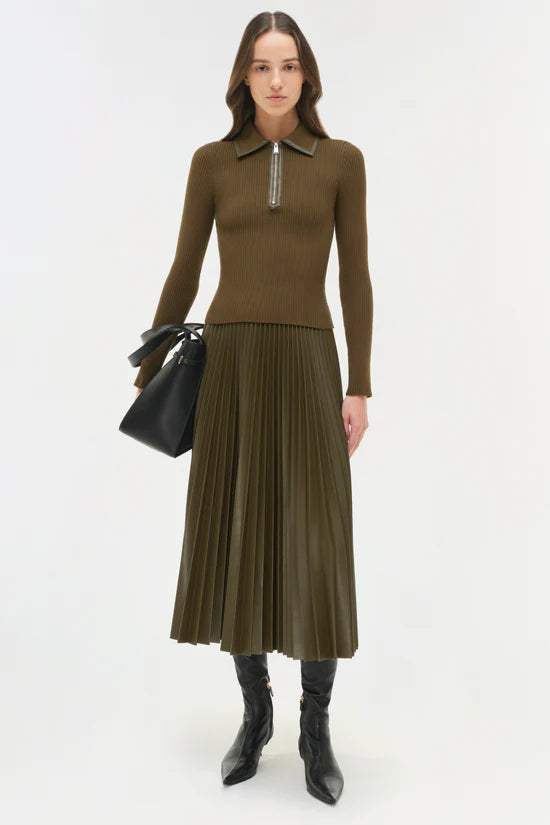 Woman wearing a brown/green sweater and pleated skirt with a black bag on a white background