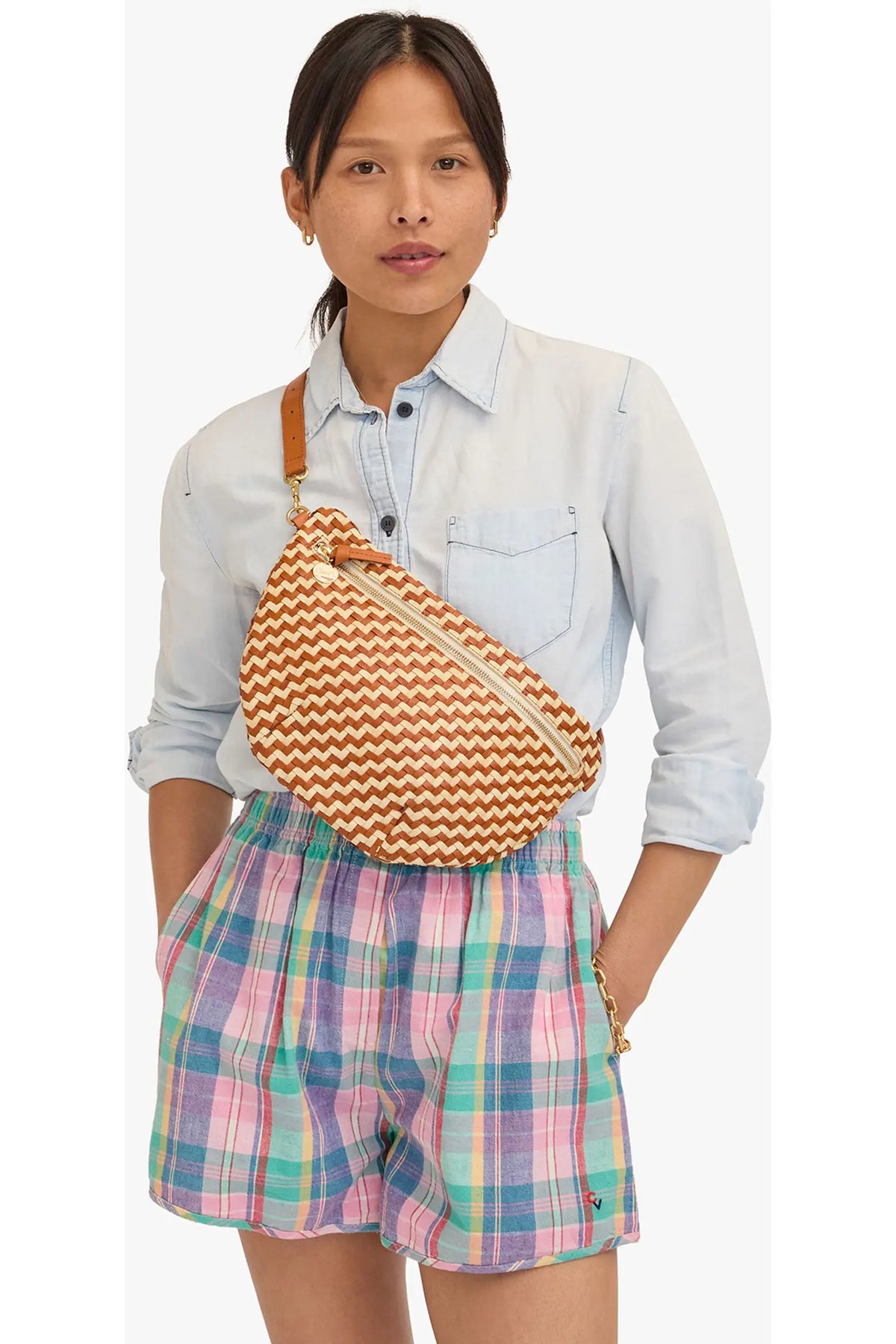 A woman wearing a light blue denim shirt and pastel plaid shorts, carrying a brown and orange chevron-patterned woven crossbody bag with a tan strap.