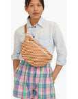A woman wearing a light blue denim shirt and pastel plaid shorts, carrying a brown and orange chevron-patterned woven crossbody bag with a tan strap.