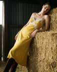 A woman leans against a large hay bale in a barn, wearing a mustard yellow midi slip dress with white lace trim at the bust and sleeves, paired with dark brown boots.