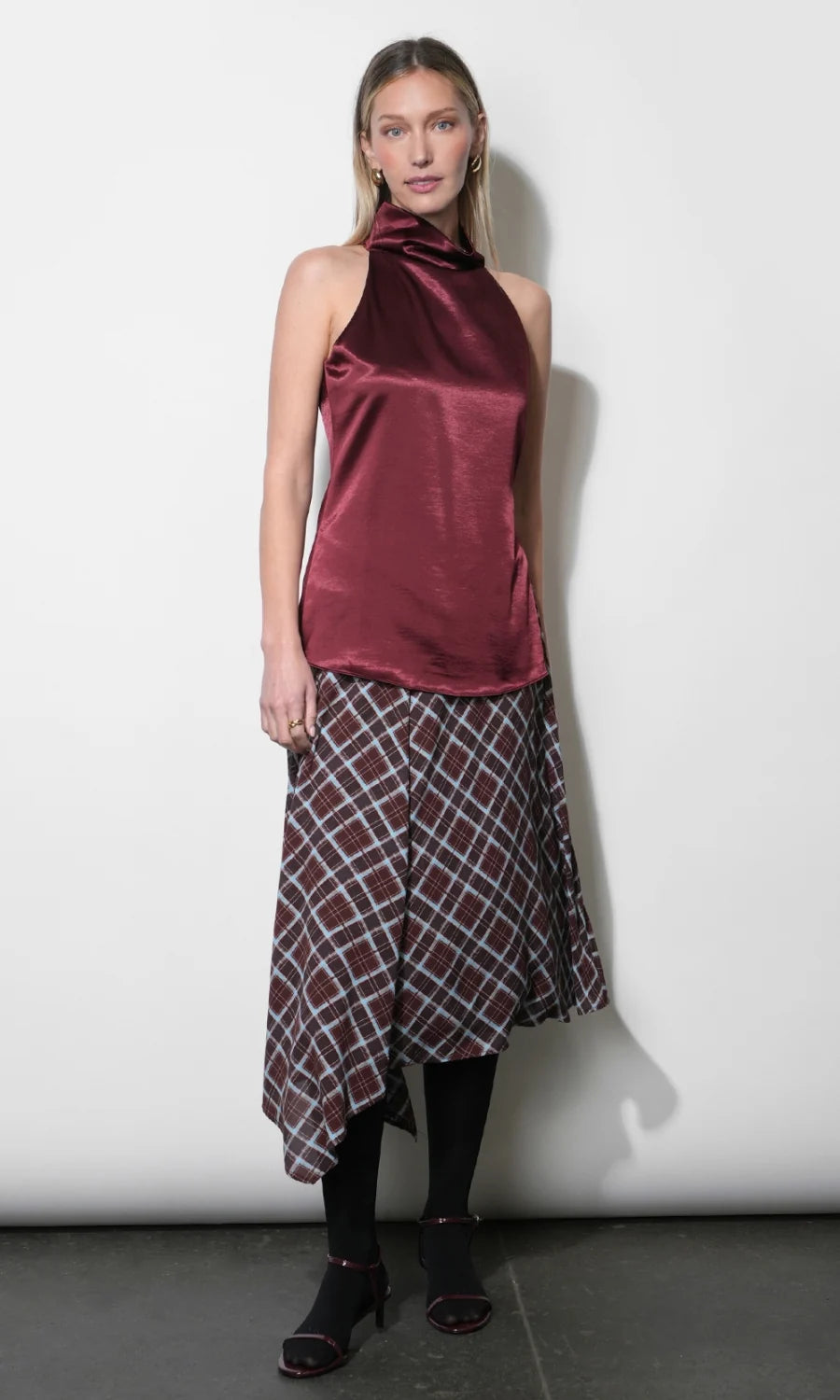 A model stands against a plain white studio wall wearing a sleeveless satin halter top in deep burgundy with a matching plaid midi skirt, black tights, and black heeled sandals.
