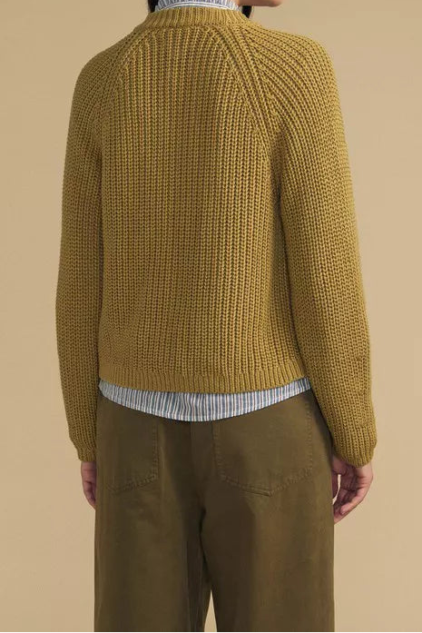 A woman shown from the back wearing a textured mustard yellow knit sweater over a striped shirt and olive green trousers, set against a solid beige background.