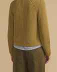 A woman shown from the back wearing a textured mustard yellow knit sweater over a striped shirt and olive green trousers, set against a solid beige background.