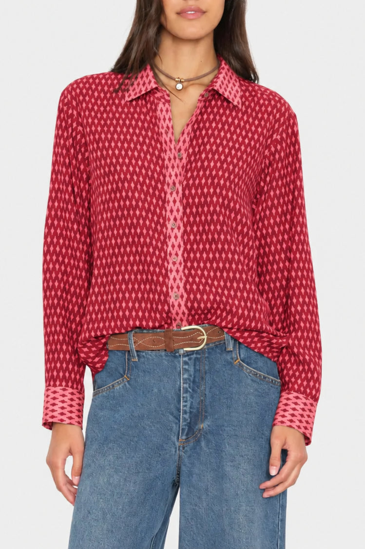 A woman wearing a red long-sleeve button-up shirt with a repeating white geometric pattern, tucked into blue jeans with a brown belt.