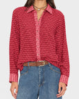 A woman wearing a red long-sleeve button-up shirt with a repeating white geometric pattern, tucked into blue jeans with a brown belt.