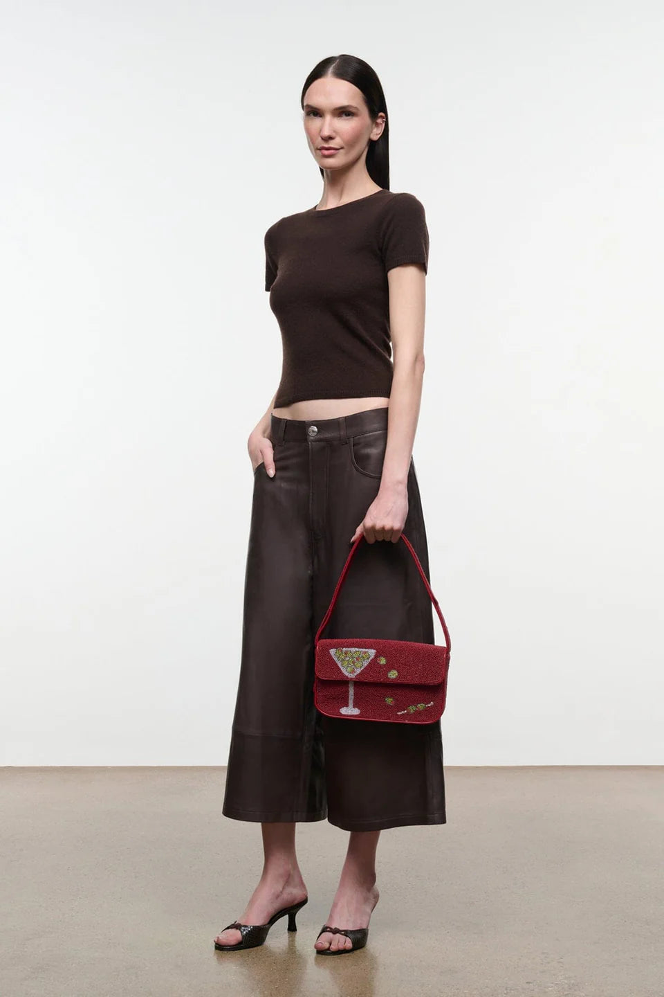 A model stands in a studio wearing a dark brown short-sleeve crop top, dark brown wide-leg cropped pants, and black heeled sandals, while holding a dark red handbag with a cocktail glass detail.