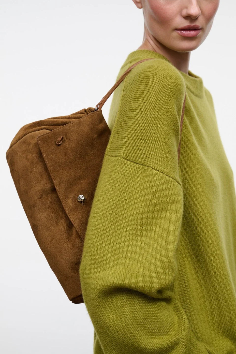 A woman wearing an oversized olive green knit sweater carries a brown suede shoulder bag on her back against a plain light gray studio background.