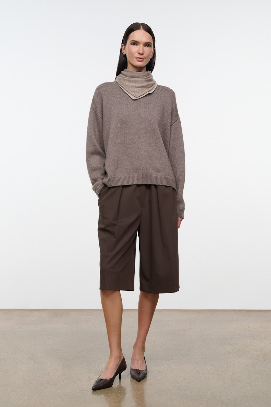 A model stands in a studio wearing a taupe-brown cowl-neck knit sweater, dark brown wide-leg cropped pants, and black pointed-toe heels against a light gray wall and concrete floor.