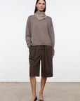 A model stands in a studio wearing a taupe-brown cowl-neck knit sweater, dark brown wide-leg cropped pants, and black pointed-toe heels against a light gray wall and concrete floor.