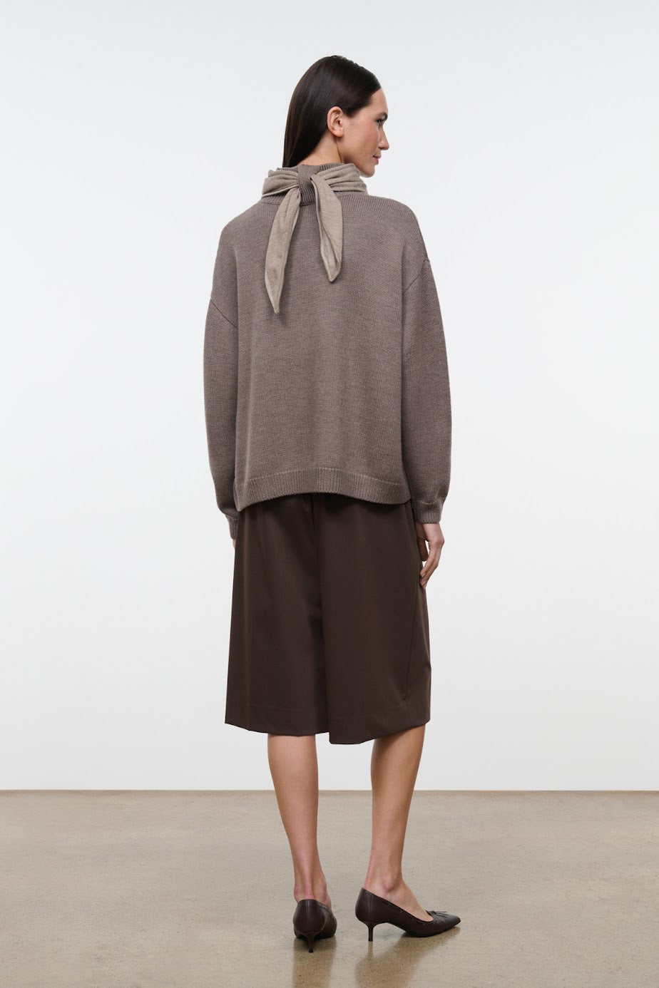 A woman stands in profile wearing a taupe knit sweater with a tied neck scarf detail, paired with dark brown midi skirt and black pointed-toe heels, against a plain white studio background.