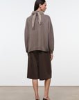 A woman stands in profile wearing a taupe knit sweater with a tied neck scarf detail, paired with dark brown midi skirt and black pointed-toe heels, against a plain white studio background.