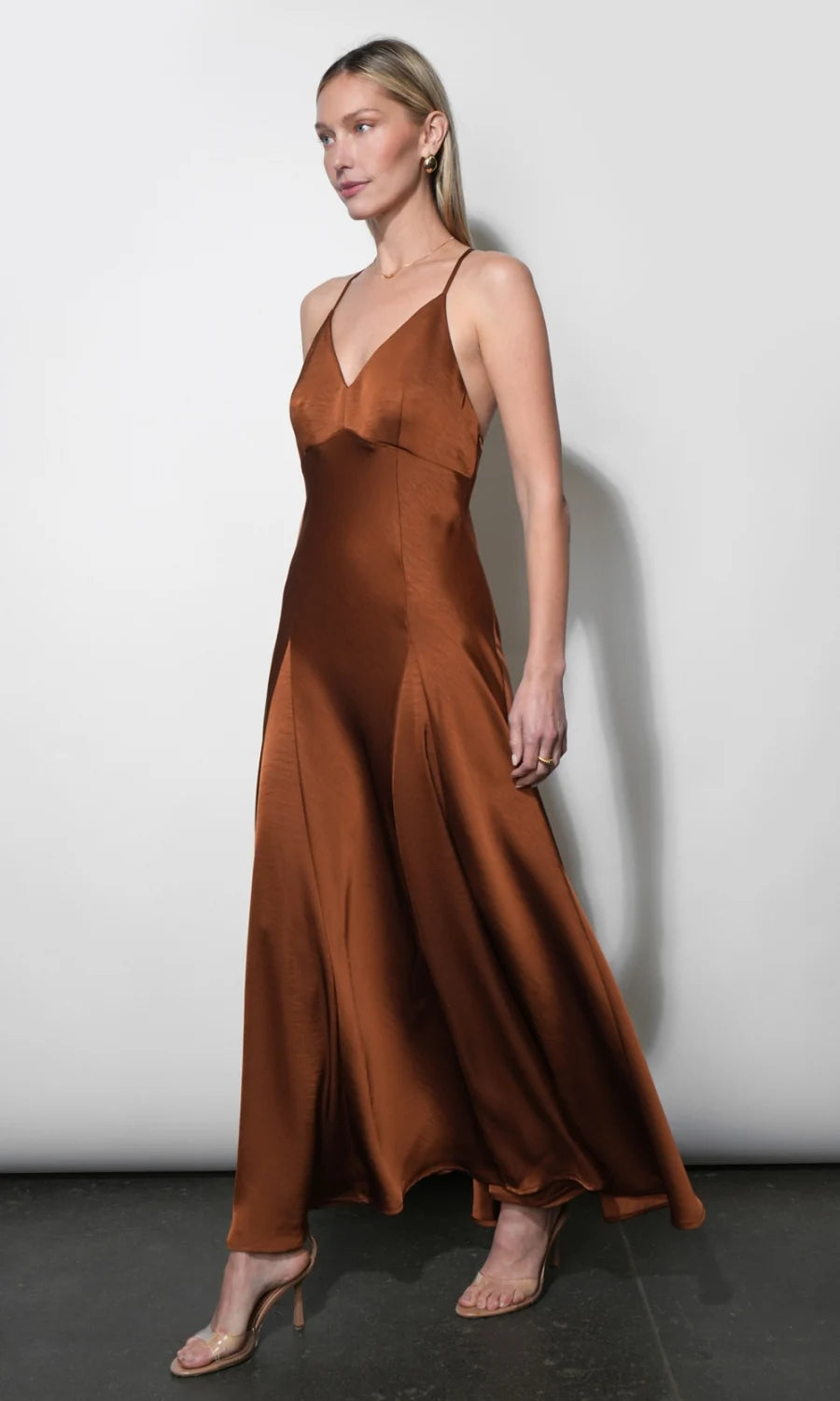 A woman modeling a floor-length satin slip dress in a warm rust brown color, standing against a plain light gray wall.