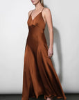 A woman modeling a floor-length satin slip dress in a warm rust brown color, standing against a plain light gray wall.
