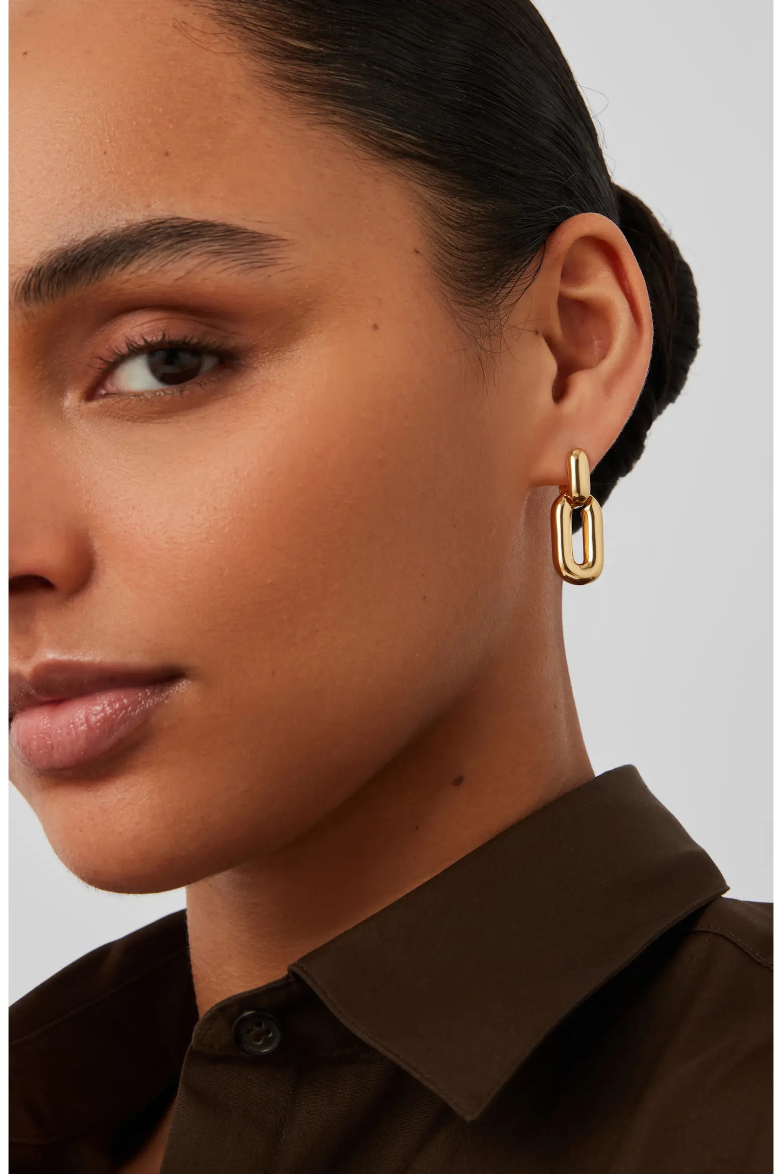 A close-up of a model wearing a gold chain-link drop earring, showing the earring against her ear. She is wearing a dark brown collared shirt against a light gray background.