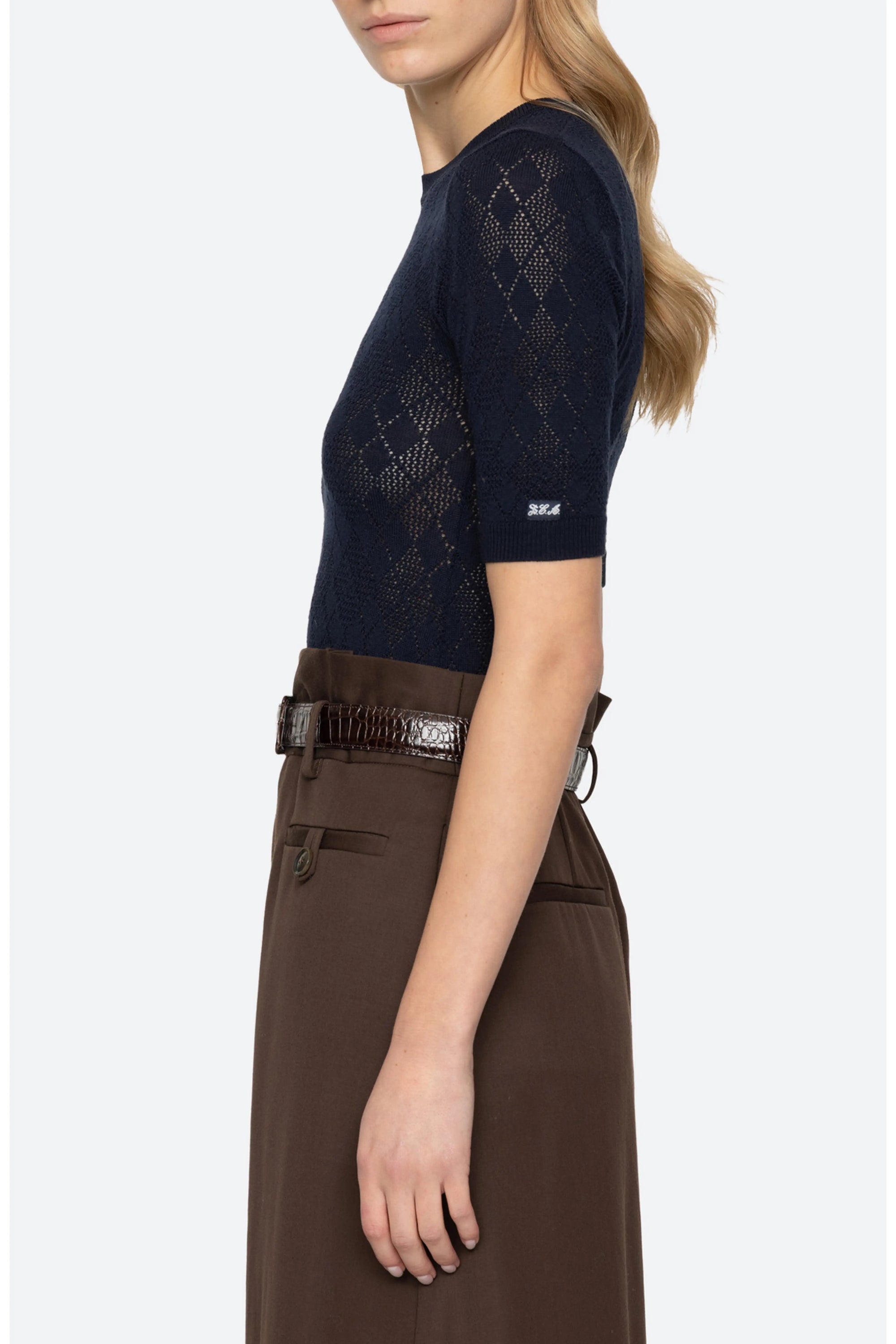 A woman shown from the side wearing a navy blue short-sleeve knit top with a textured argyle pattern and a small white logo on the sleeve, paired with a brown high-waisted skirt.