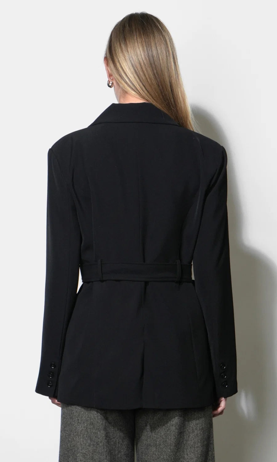 A woman shown from the back wearing a black belted blazer and gray trousers against a plain light background.