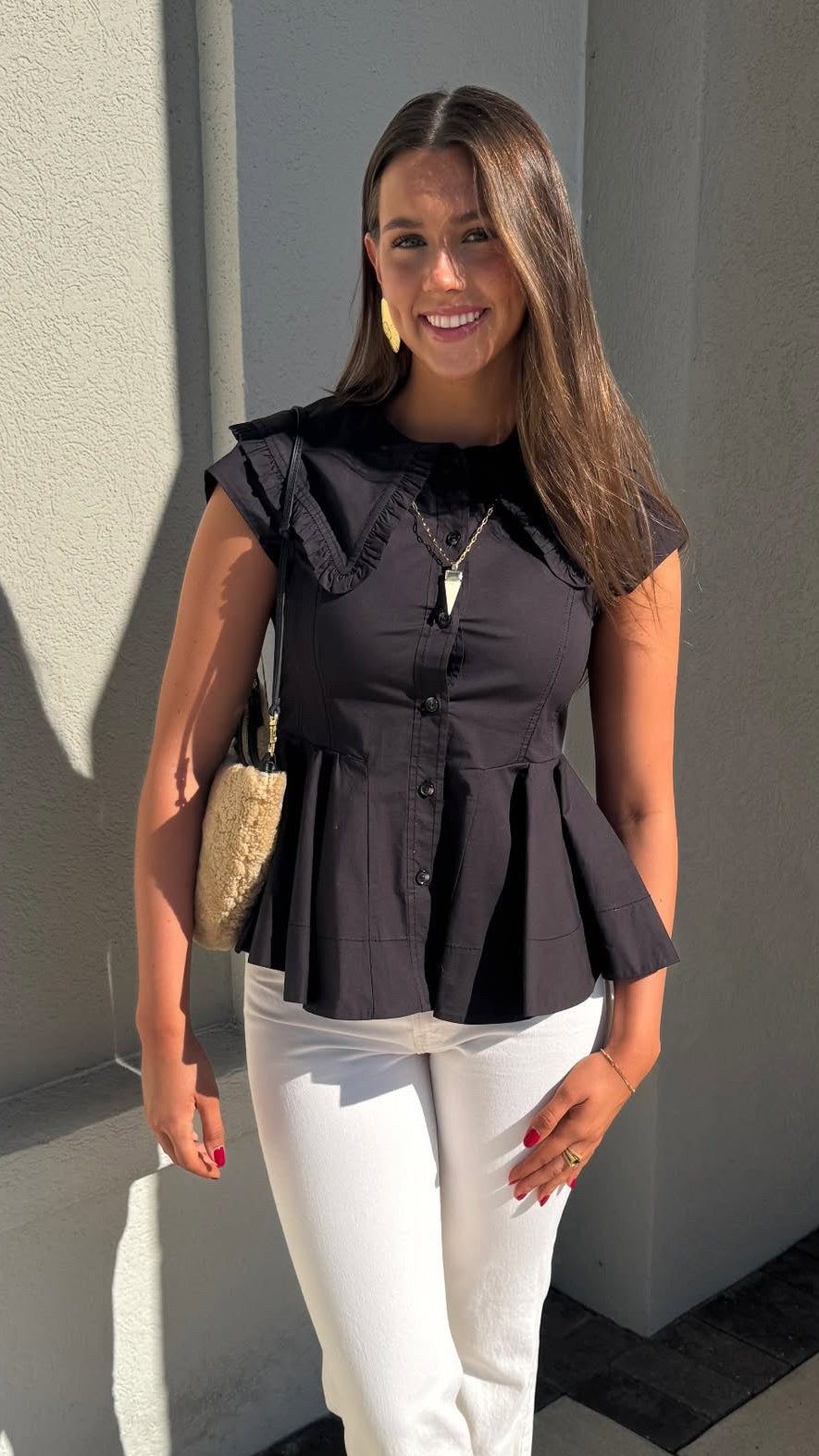 Woman wearing a black peplum top and white pants standing against a gray wall.