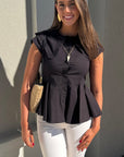 Woman wearing a black peplum top and white pants standing against a gray wall.