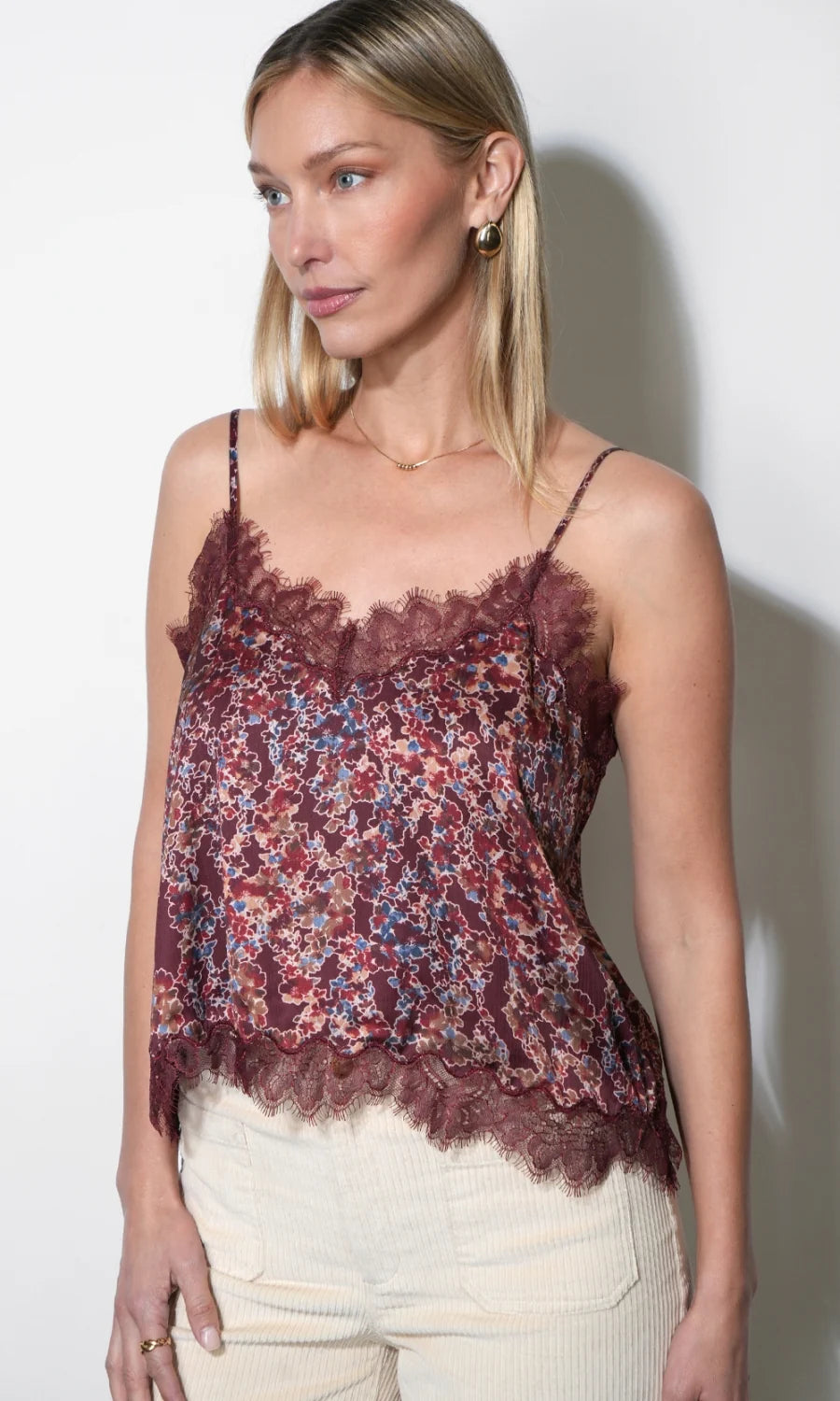 A woman modeling a burgundy floral lace camisole top with thin straps and a ruffled hem, paired with cream pants, against a plain white wall.