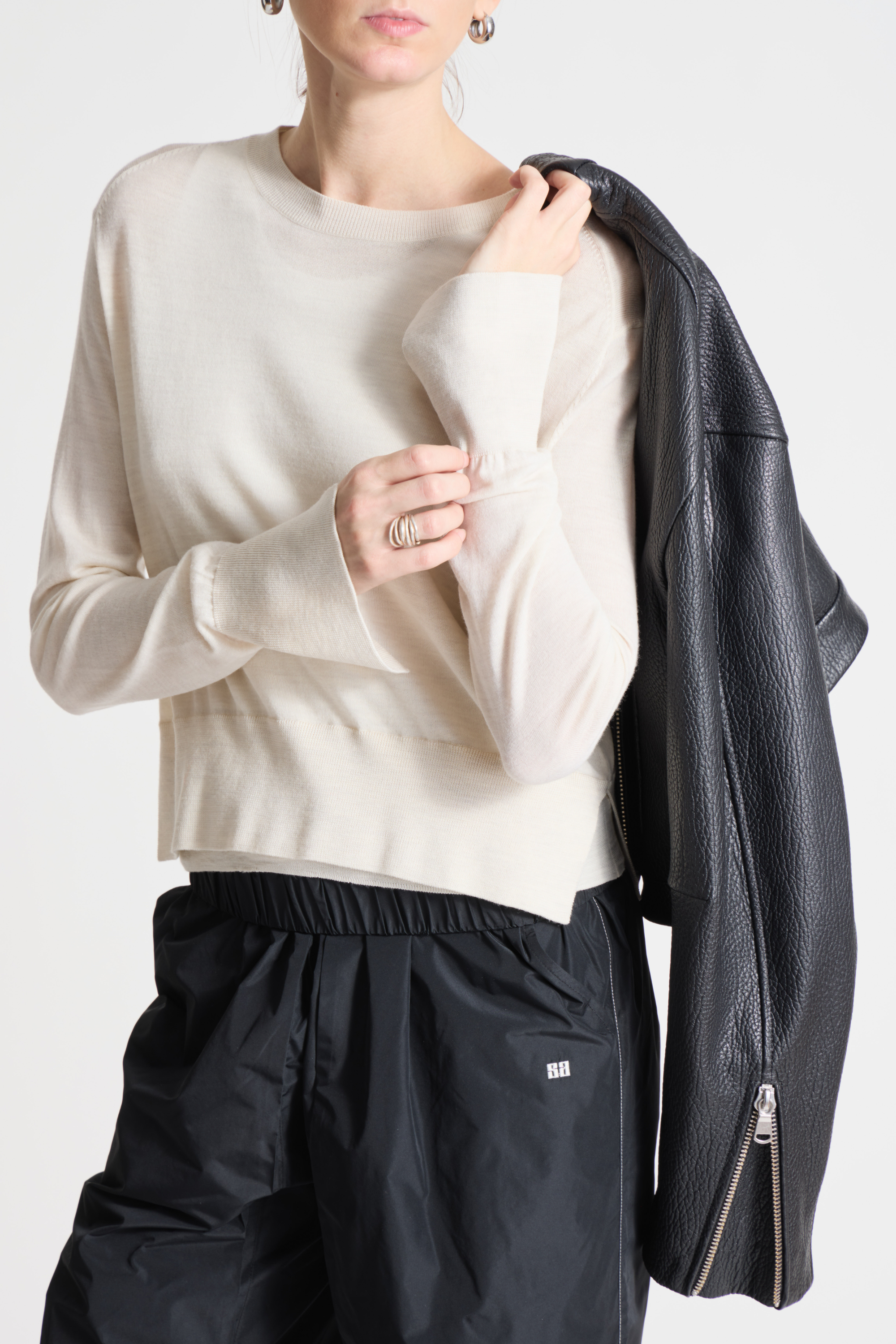 Person wearing a beige sweater and black pants holding a black leather jacket.