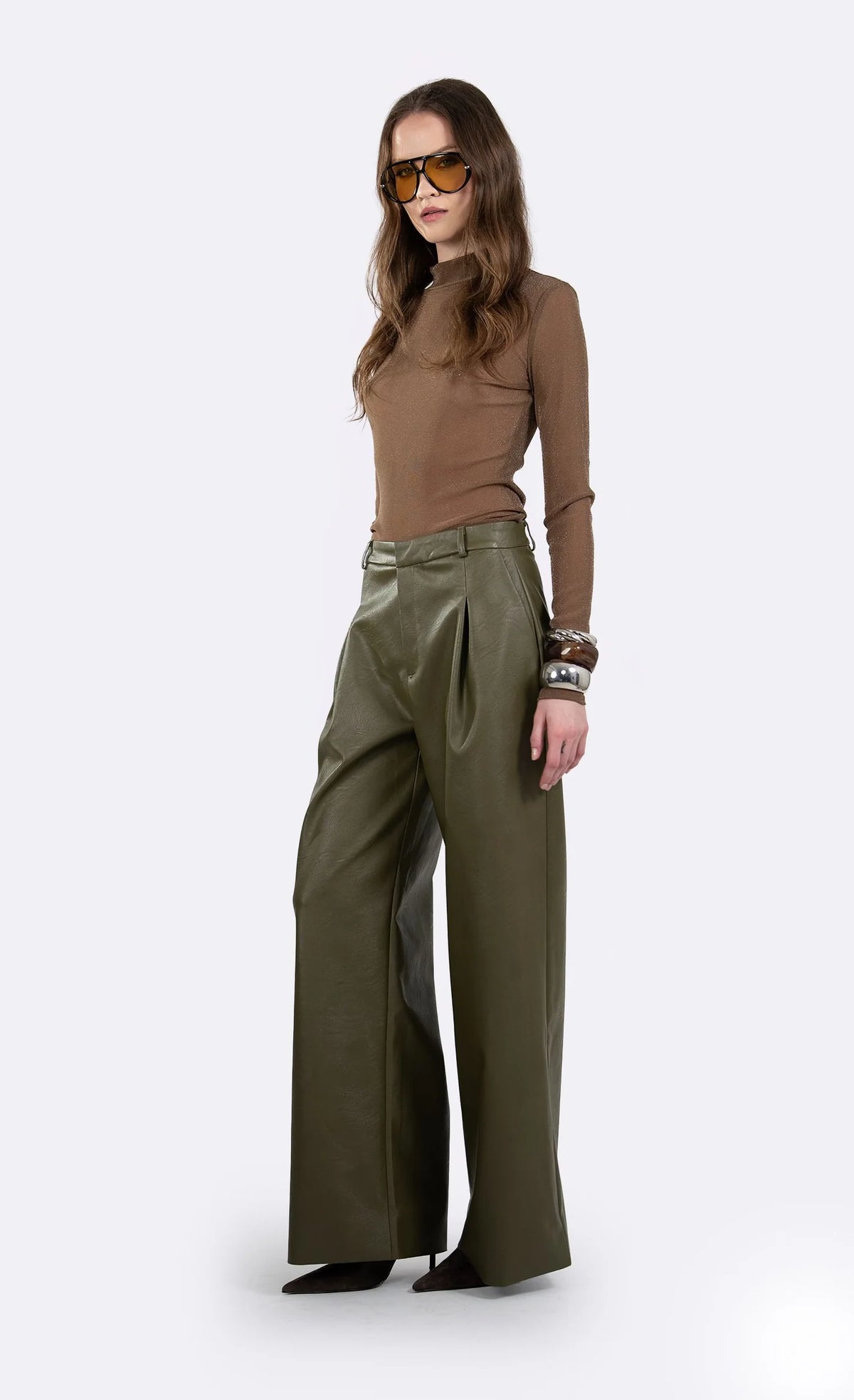 A model wearing a brown mock-neck long-sleeve top and olive green wide-leg trousers, styled with brown sunglasses and bracelets, standing against a light gray studio background.