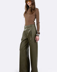 A model wearing a brown mock-neck long-sleeve top and olive green wide-leg trousers, styled with brown sunglasses and bracelets, standing against a light gray studio background.