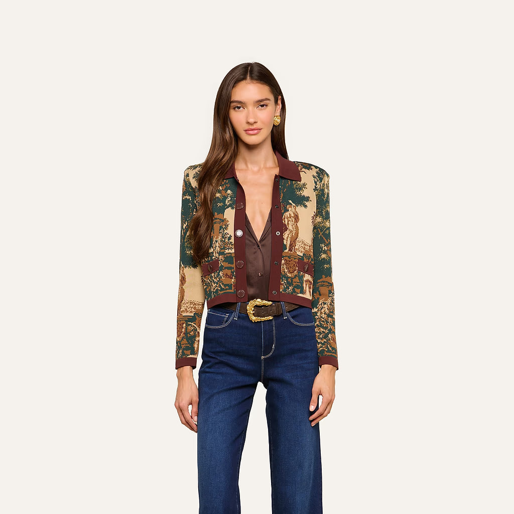 A model wearing a cropped, long-sleeve button-front cardigan with a detailed landscape tapestry print in beige, green, and brown, paired with dark blue jeans and a gold-buckled belt, against a solid off-white background.