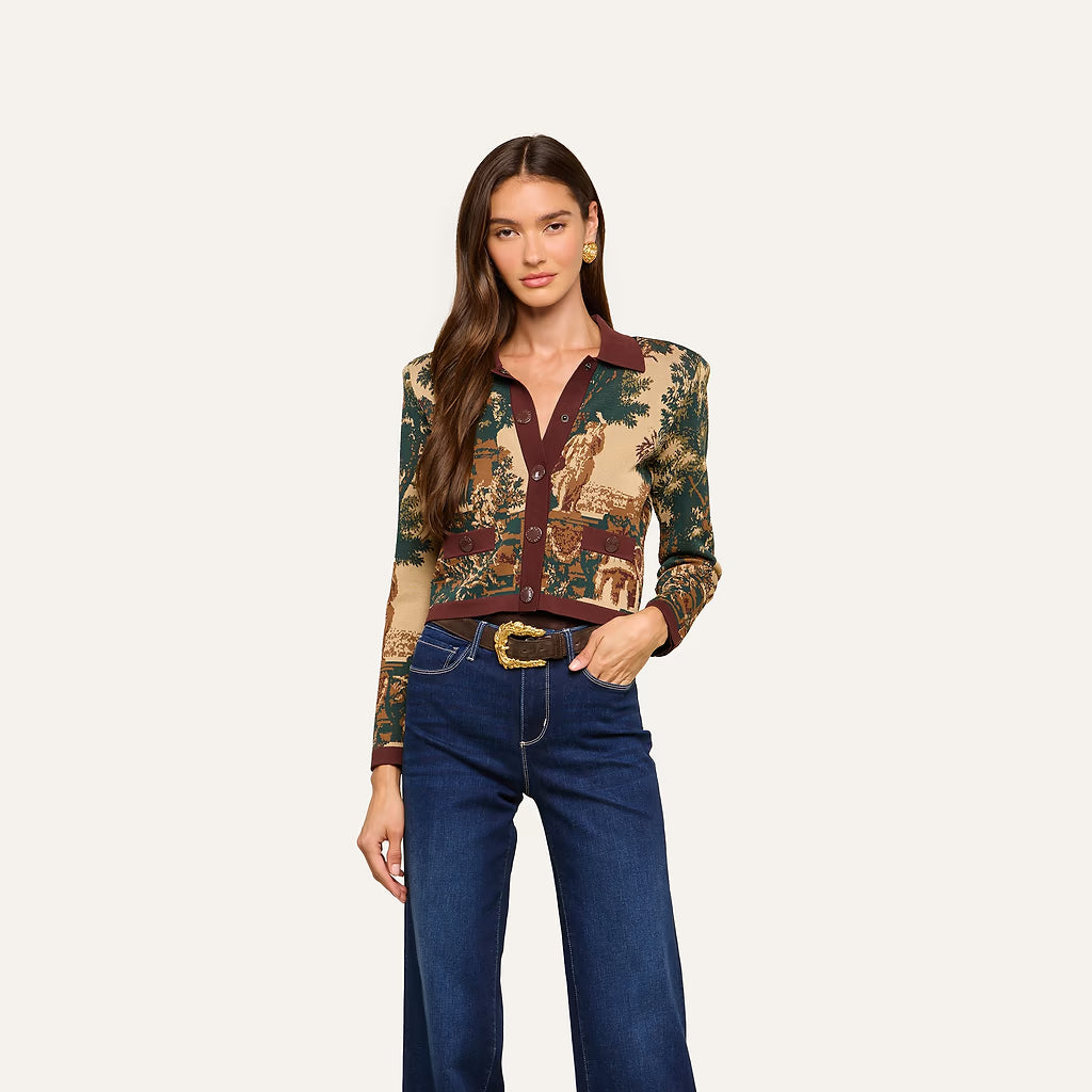 A woman modeling a long-sleeve button-up shirt with a detailed tapestry-style print in green, beige, and brown, paired with dark blue jeans and a gold-buckled belt, against a solid off-white background.