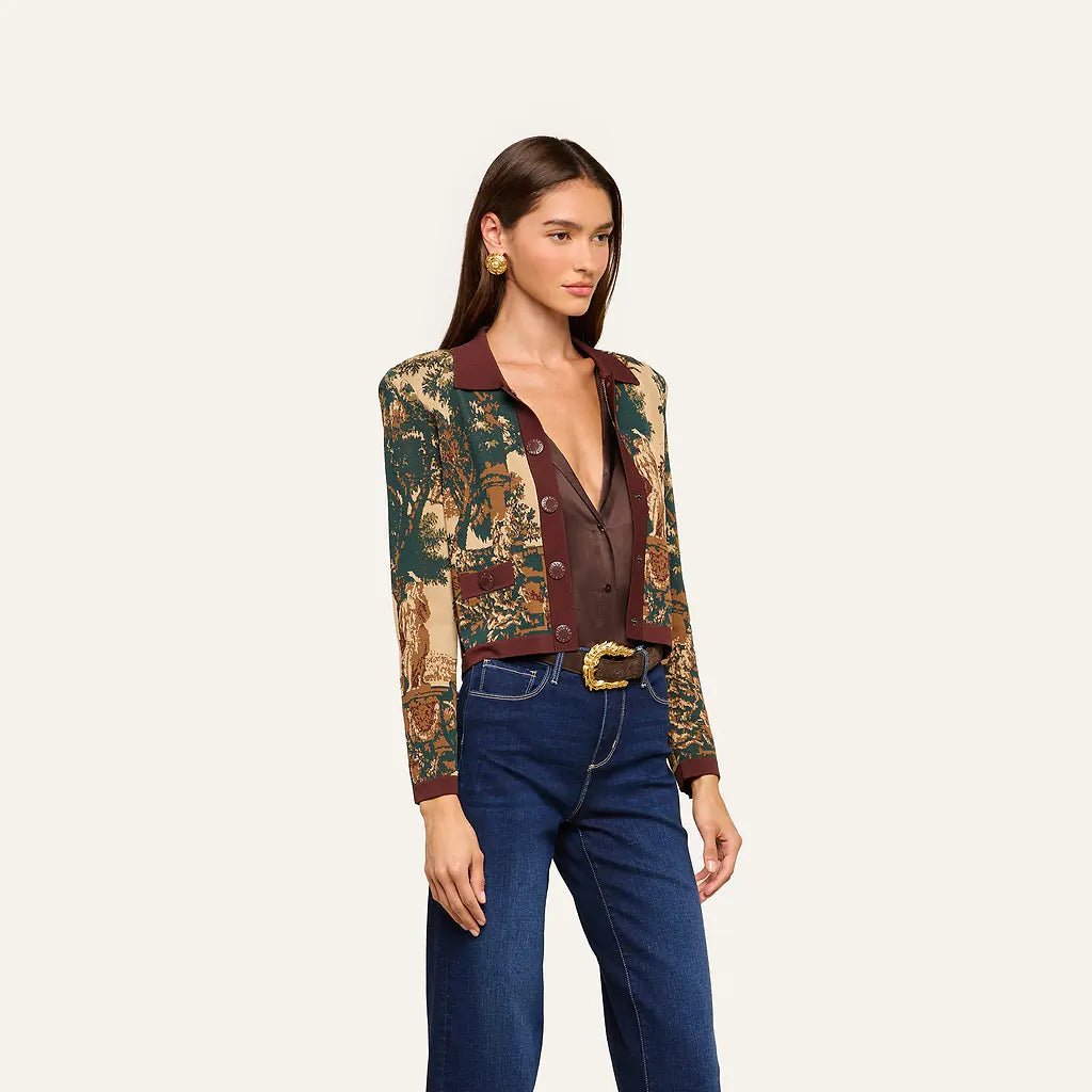 A model wearing a cropped, long-sleeve button-front jacket with a brown and green landscape print on the sleeves and a solid dark brown collar and front placket, paired with dark blue jeans and a gold belt, against a light beige background.