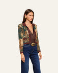 A model wearing a cropped, long-sleeve button-front jacket with a brown and green landscape print on the sleeves and a solid dark brown collar and front placket, paired with dark blue jeans and a gold belt, against a light beige background.