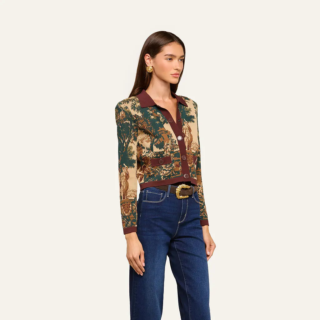 A woman modeling a long-sleeve collared button-up shirt with an intricate multicolor tapestry-style print in green, gold, and brown, paired with a matching brown belt and dark blue jeans, set against a solid off-white background.