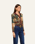 A woman modeling a long-sleeve collared button-up shirt with an intricate multicolor tapestry-style print in green, gold, and brown, paired with a matching brown belt and dark blue jeans, set against a solid off-white background.