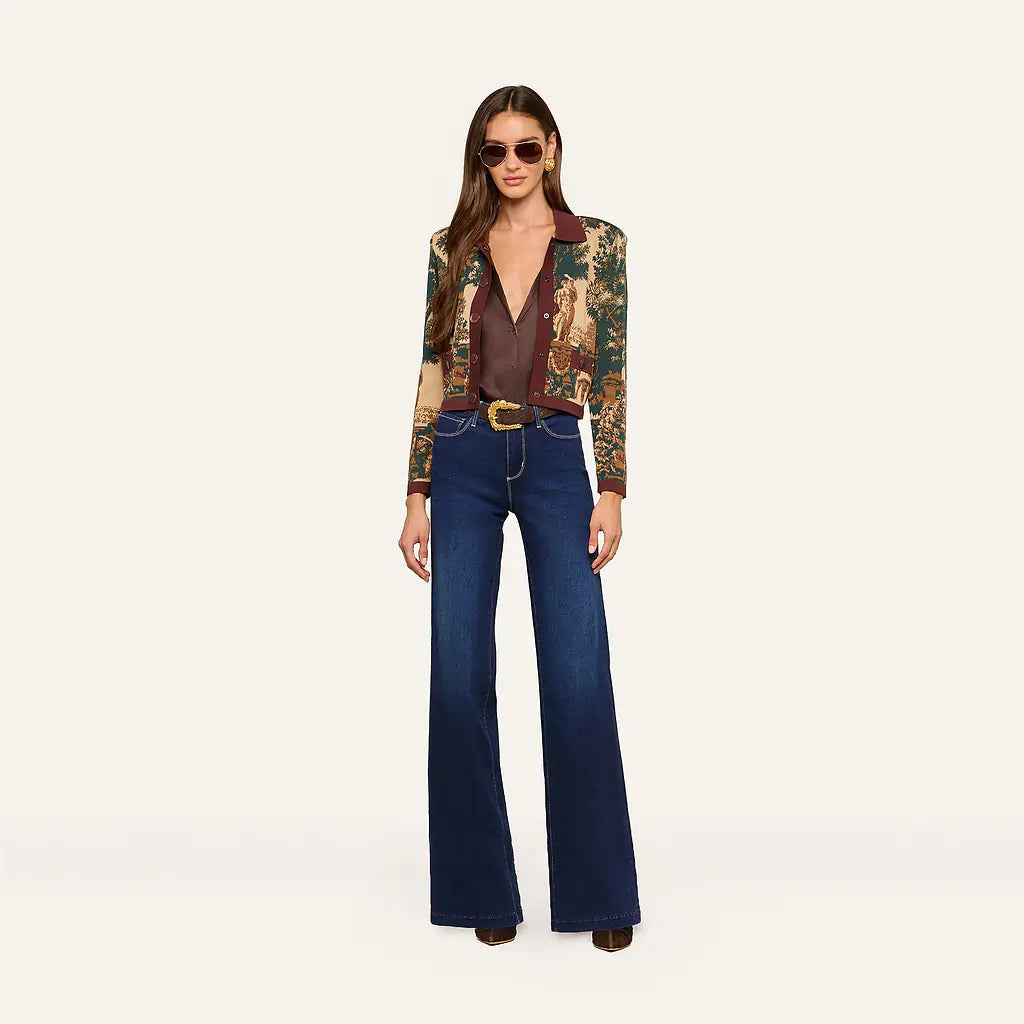 A model stands against a plain light background wearing a patterned long-sleeve blazer with a brown shirt, dark blue wide-leg jeans, and sunglasses.