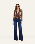 A model stands against a plain light background wearing a patterned long-sleeve blazer with a brown shirt, dark blue wide-leg jeans, and sunglasses.