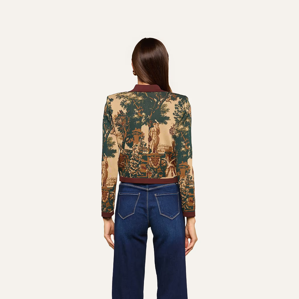 A woman viewed from the back wearing a cropped long-sleeve bomber jacket with a detailed classical landscape toile pattern in beige, green, and brown, paired with dark blue jeans, against a solid off-white background.