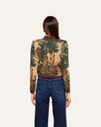 A woman viewed from the back wearing a cropped long-sleeve bomber jacket with a detailed classical landscape toile pattern in beige, green, and brown, paired with dark blue jeans, against a solid off-white background.