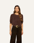 A woman modeling a short-sleeve crewneck knit sweater in dark brown, paired with dark brown wide-leg trousers and a textured belt, against a solid off-white background.