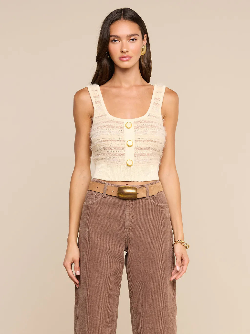 A woman modeling a cream-colored textured knit tank top with three gold buttons, paired with brown wide-leg jeans and a brown belt, against a solid beige background.