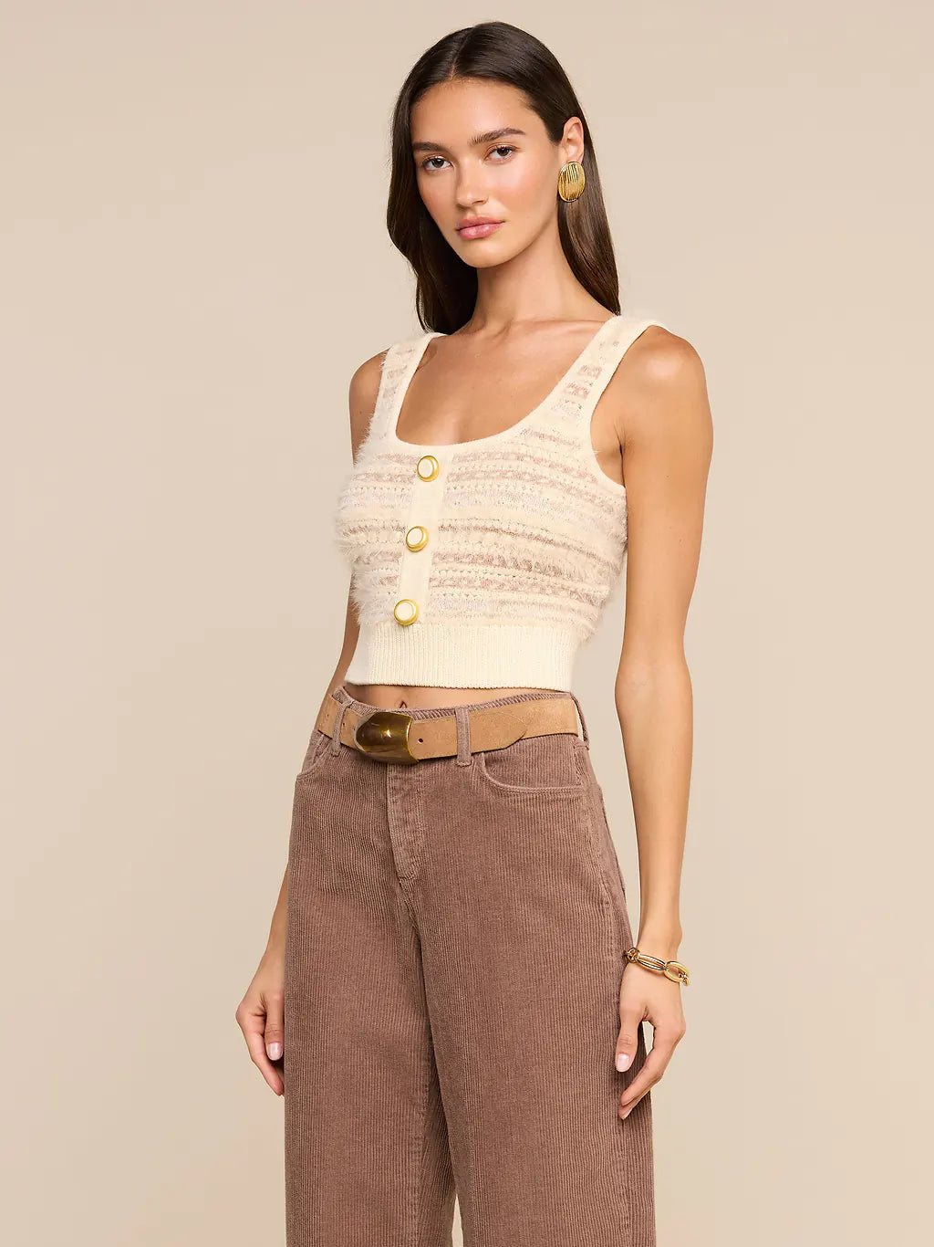 A woman modeling a cream knit cropped tank top with gold buttons, paired with brown wide-leg pants and a tan belt, against a solid beige background.