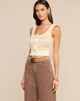 A woman modeling a cream knit cropped tank top with gold buttons, paired with brown wide-leg pants and a tan belt, against a solid beige background.