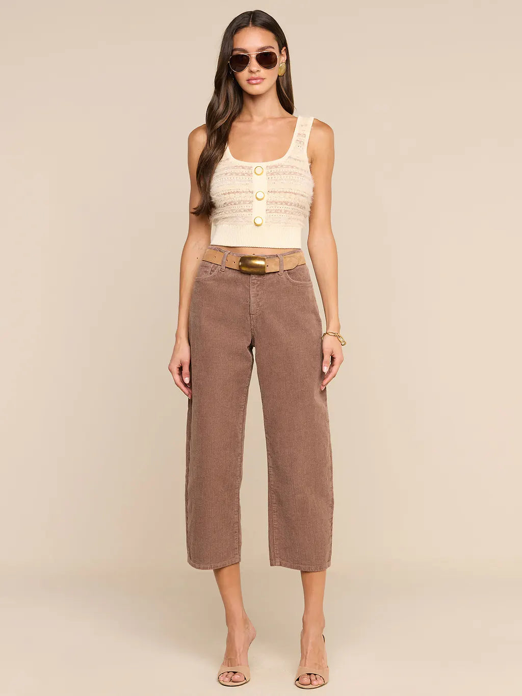 A woman modeling a cream ribbed knit cropped tank top with gold buttons and brown wide-leg cropped pants with a tan belt, set against a solid beige studio background.