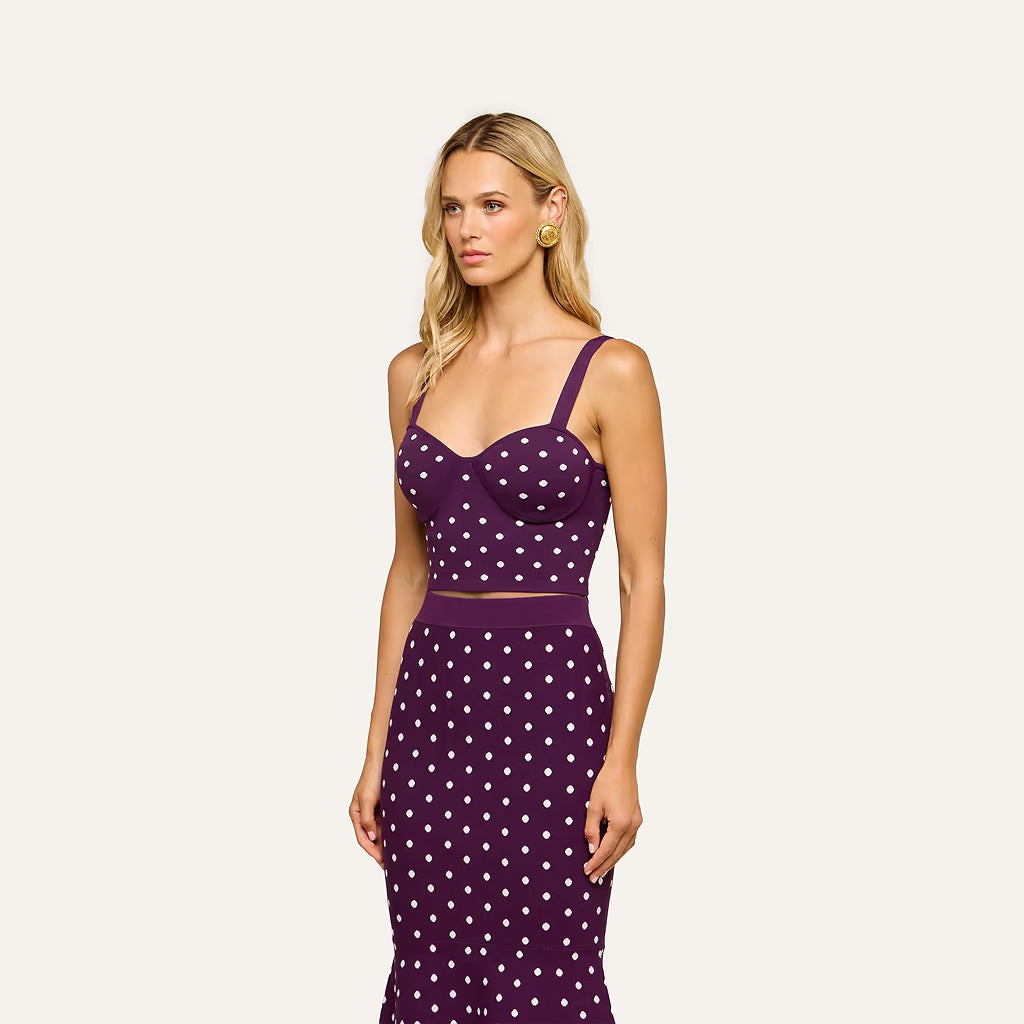 A woman modeling a two-piece sleeveless dress set in deep purple with a white polka dot pattern, featuring a structured sweetheart bustier top and a matching high-waisted skirt, set against a solid off-white background.
