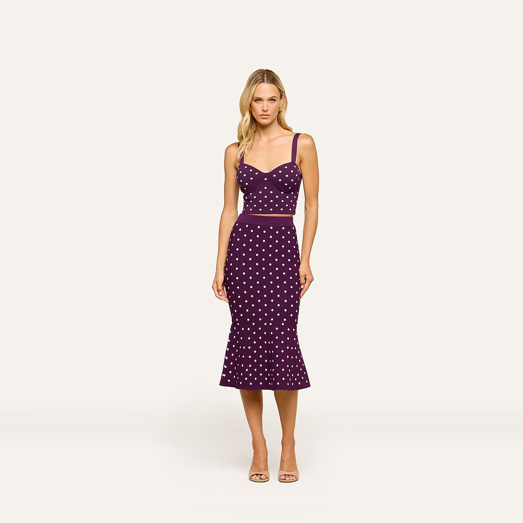 A woman modeling a sleeveless midi dress with a sweetheart neckline, thin shoulder straps, a fitted bodice, and a flared skirt. The dress is deep purple with a white polka dot pattern and is shown against a plain light gray studio background.