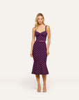 A woman modeling a sleeveless midi dress with a sweetheart neckline, thin shoulder straps, a fitted bodice, and a flared skirt. The dress is deep purple with a white polka dot pattern and is shown against a plain light gray studio background.