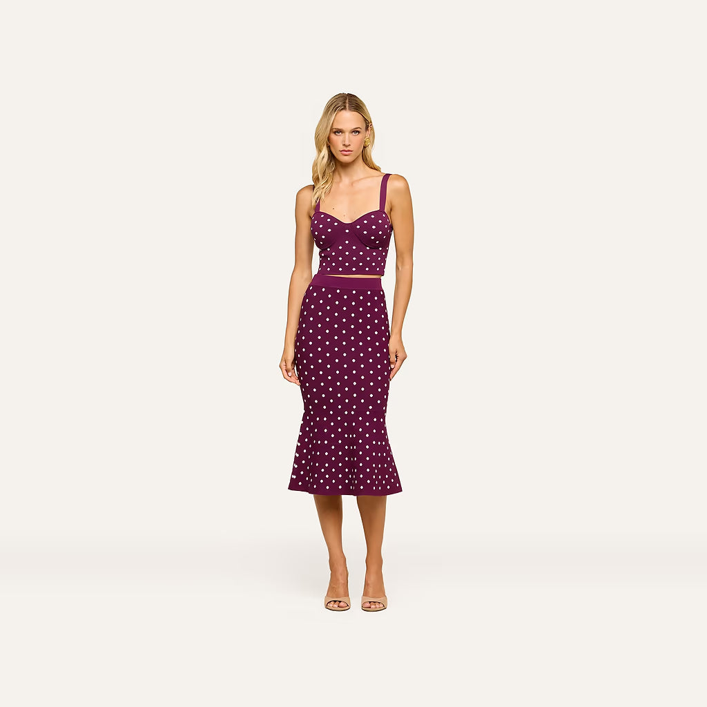 A woman modeling a sleeveless midi dress with a sweetheart neckline, thin shoulder straps, a fitted bodice, and a flared skirt. The dress is deep purple with a white polka-dot pattern and a thin purple waistband.