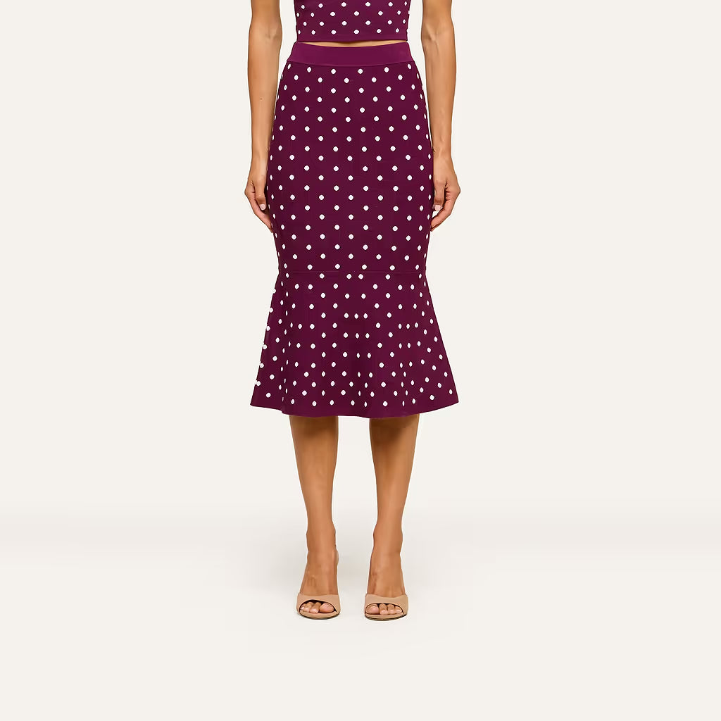 A model wearing a dark purple midi skirt with a white polka dot pattern, paired with a matching crop top and beige heeled sandals, set against a plain light background.