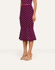 A model wearing a dark purple midi skirt with white polka dots and beige heeled sandals, shown from the waist down against a light off-white background.